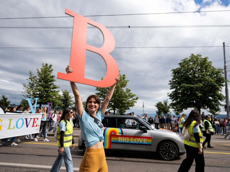 Une femme porte un grand B en se déplaçant devant la MINI avec l’arc-en-ciel et l’inscription BIG LOVE